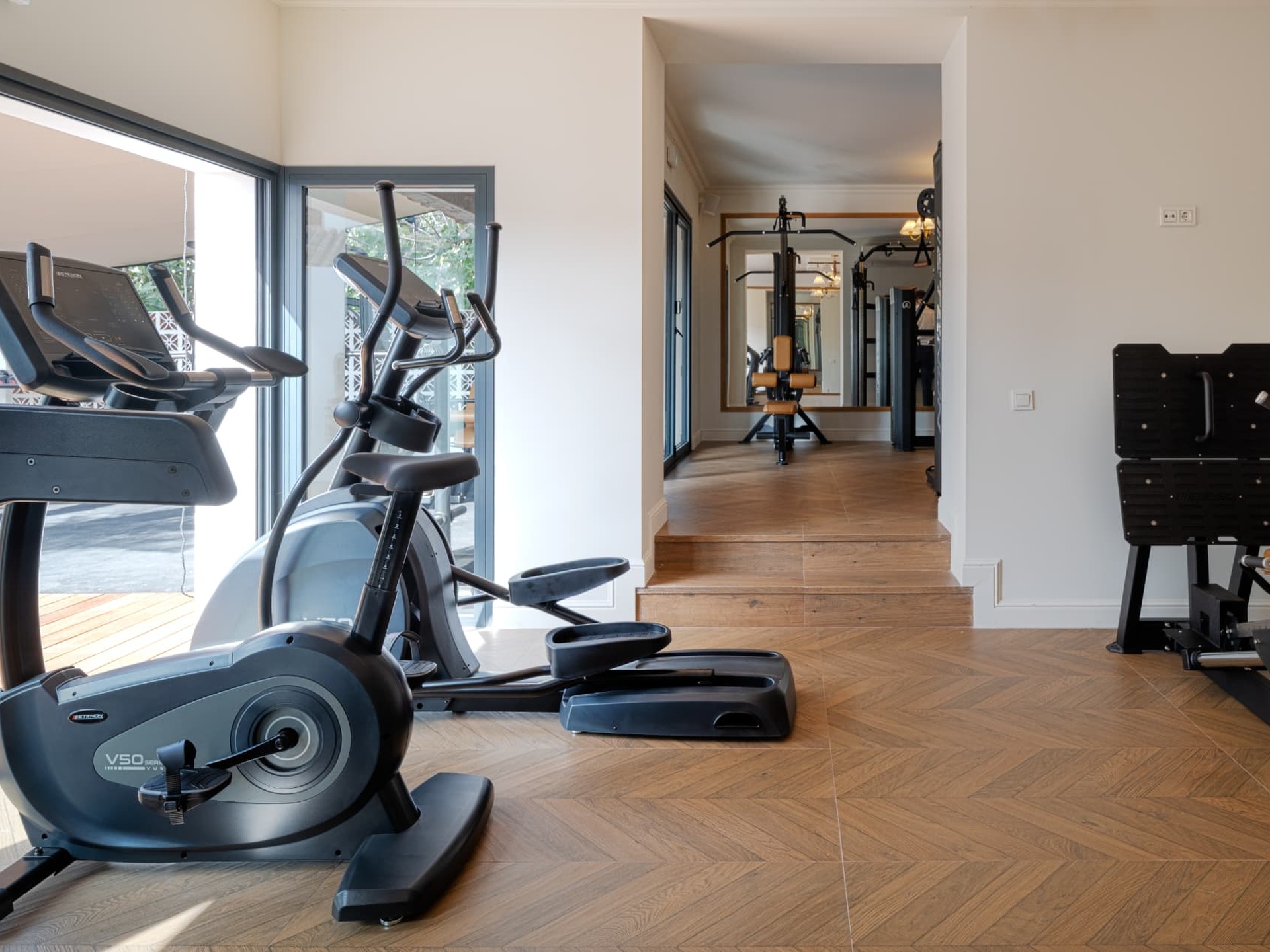Bright hotel gym interior with cardio machines, herringbone wood flooring and floor-to-ceiling glazing opening to a terrace.