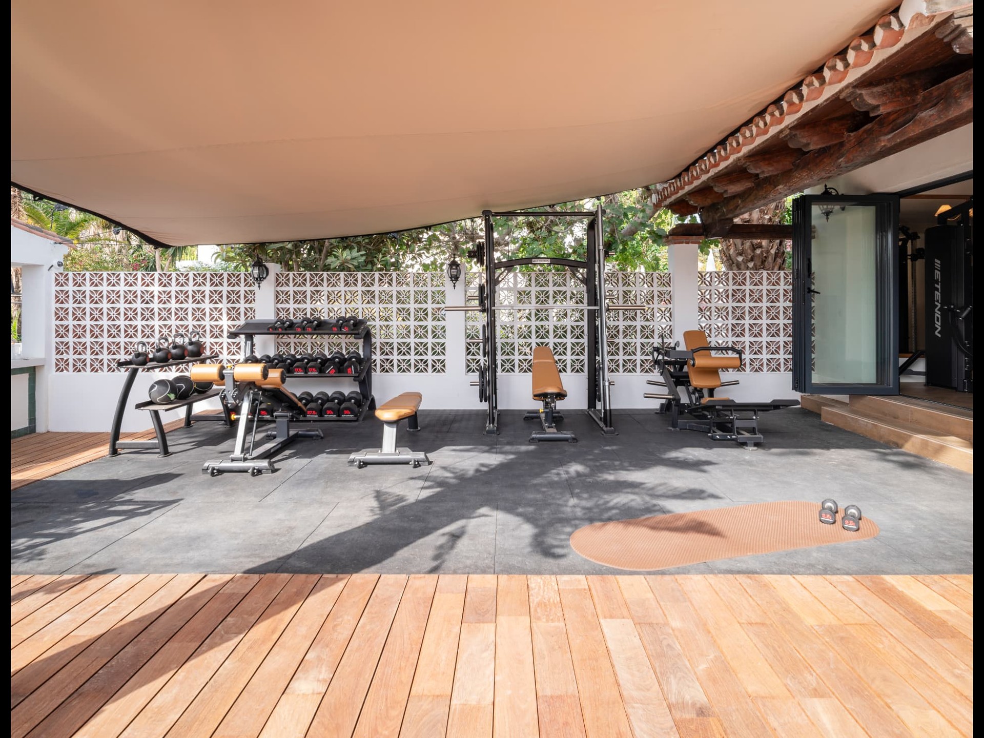 Covered outdoor hotel gym terrace with strength equipment, camel leather benches, white lattice screens and timber decking.