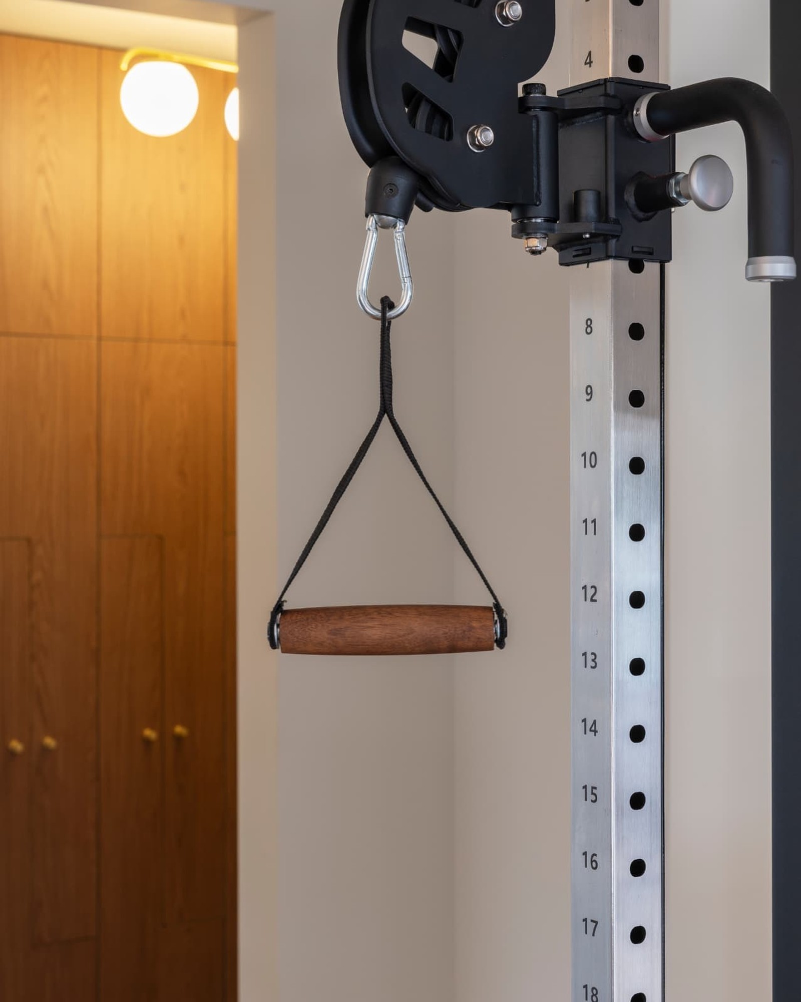 Custom cable station handle with a sculpted walnut grip and brushed steel carabiner in a warm hotel gym interior.